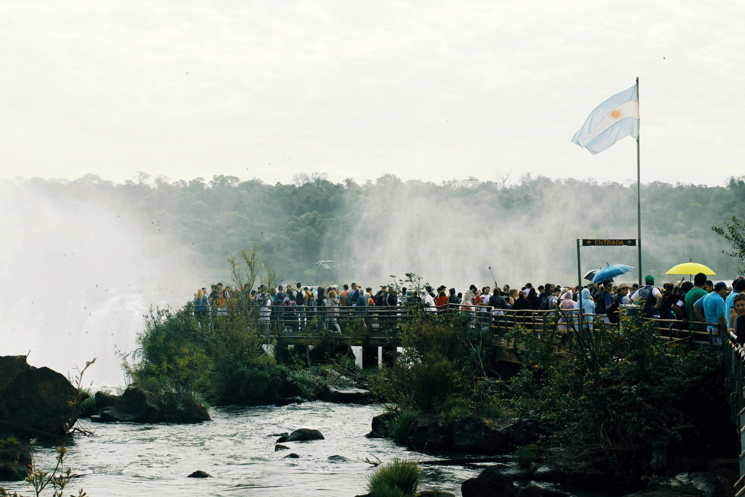 A crowded boardwalk with tourists overlooking the misty Iguazu Falls, featuring scenic views and the Argentine flag.